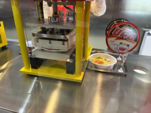 任意のかやくを入れたUFOに中蓋をする機械。待ち時間は自分が書いた外蓋を見えるように置いてくれる。