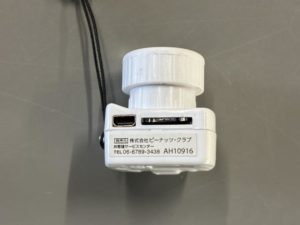 Digital Snap Camera microの下側を撮った写真。充電口（microUSB）とmicroSDを差し込むスロットがあり、その下にピーナッツクラブとその連絡先が書いてあるシールが貼ってある。